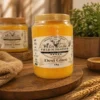 A 1kg clear glass jar of golden yellow Desi Ghee on a wooden platter, surrounded by rustic burlap and wheat stalks.