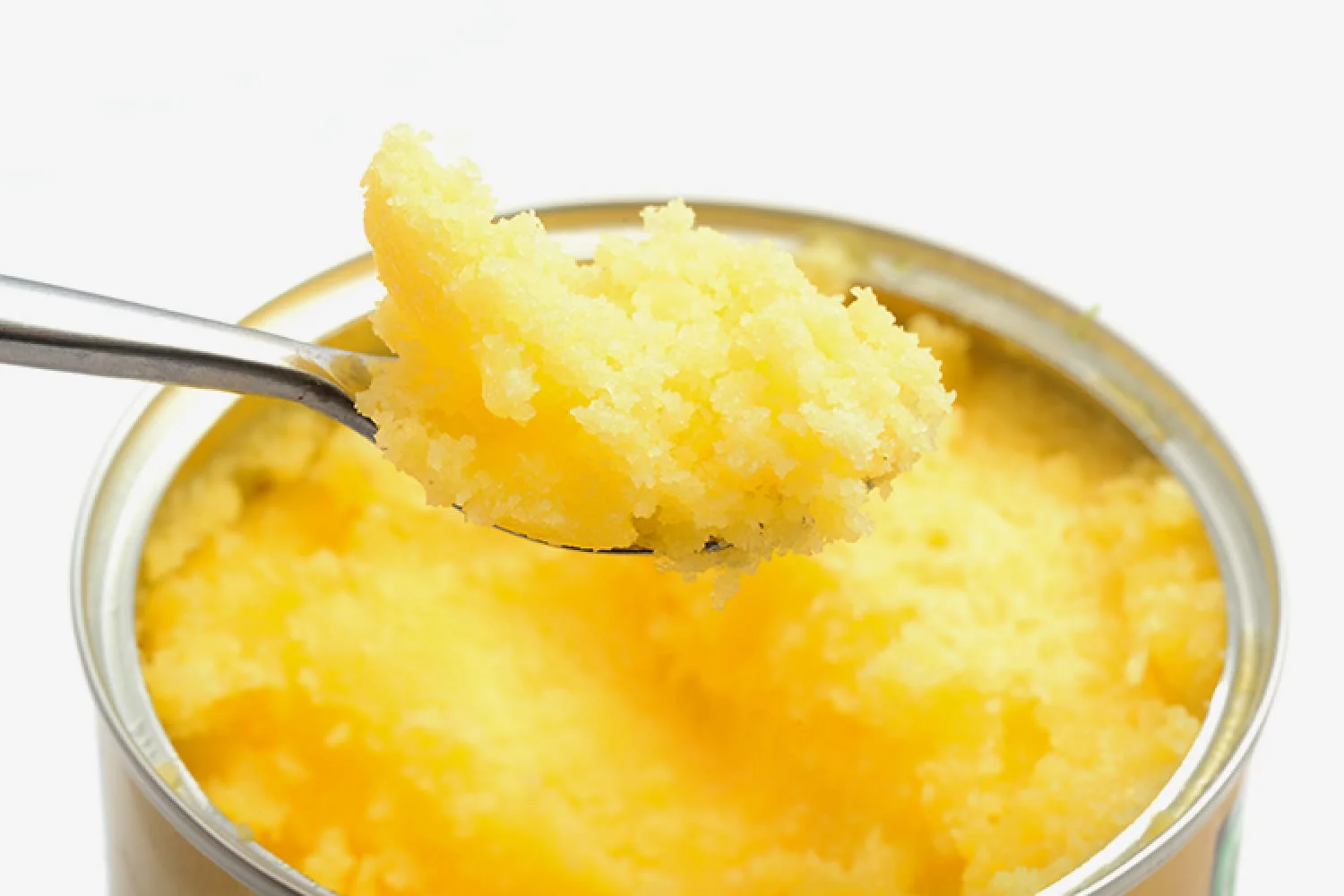A silver spoon scooping a portion of grainy, yellow pure desi cow ghee from a metal tin against a white background.